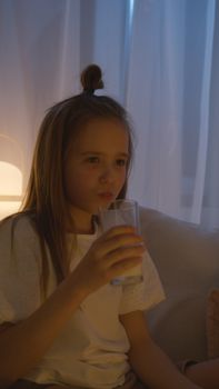 A young Caucasian girl drinks milk indoors during the evening, creating a warm and cozy atmosphere.