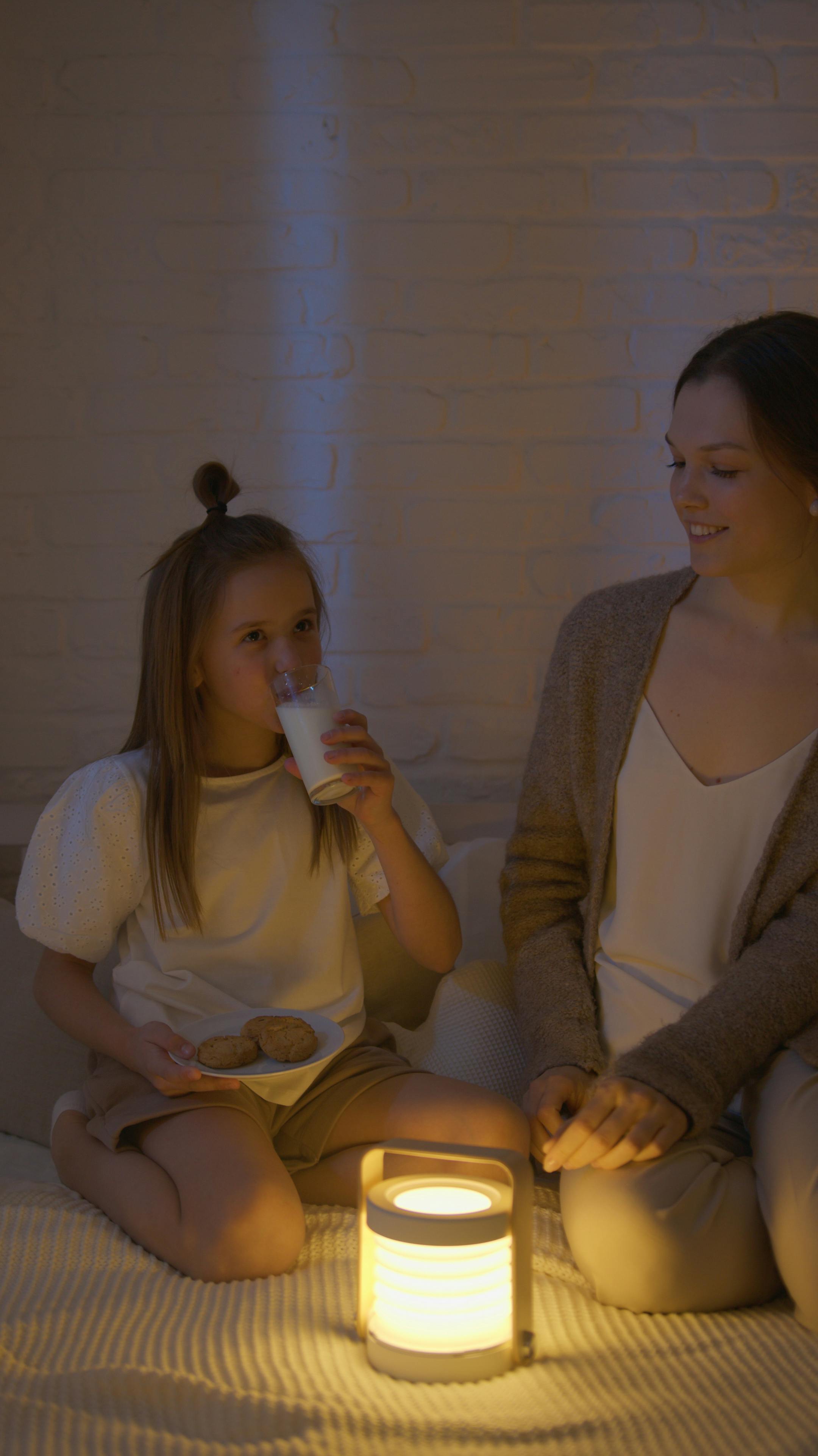 woman sitting beside a girl drinking a milk on bed