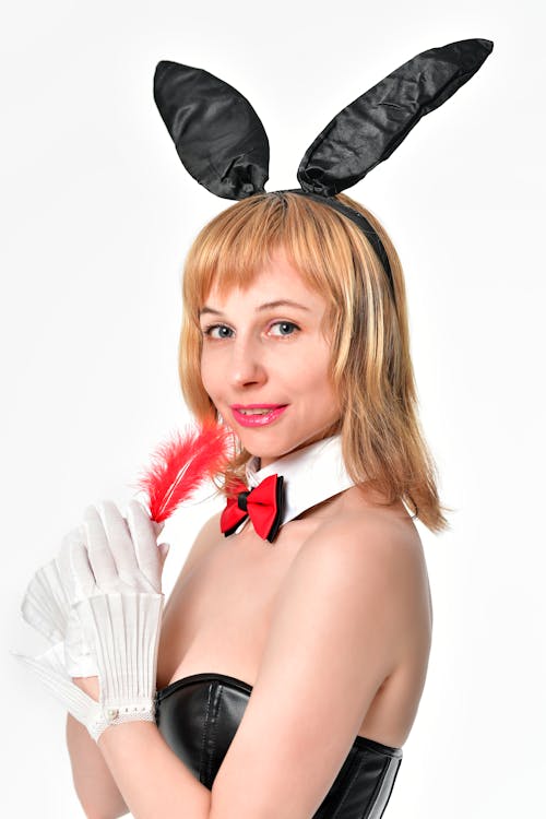 Side view of female wearing sexy black bunny ears and leather corset touching face with feather and looking at camera with smile