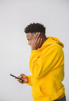 Side profile of a man in a yellow hoodie using a smartphone against a neutral background.