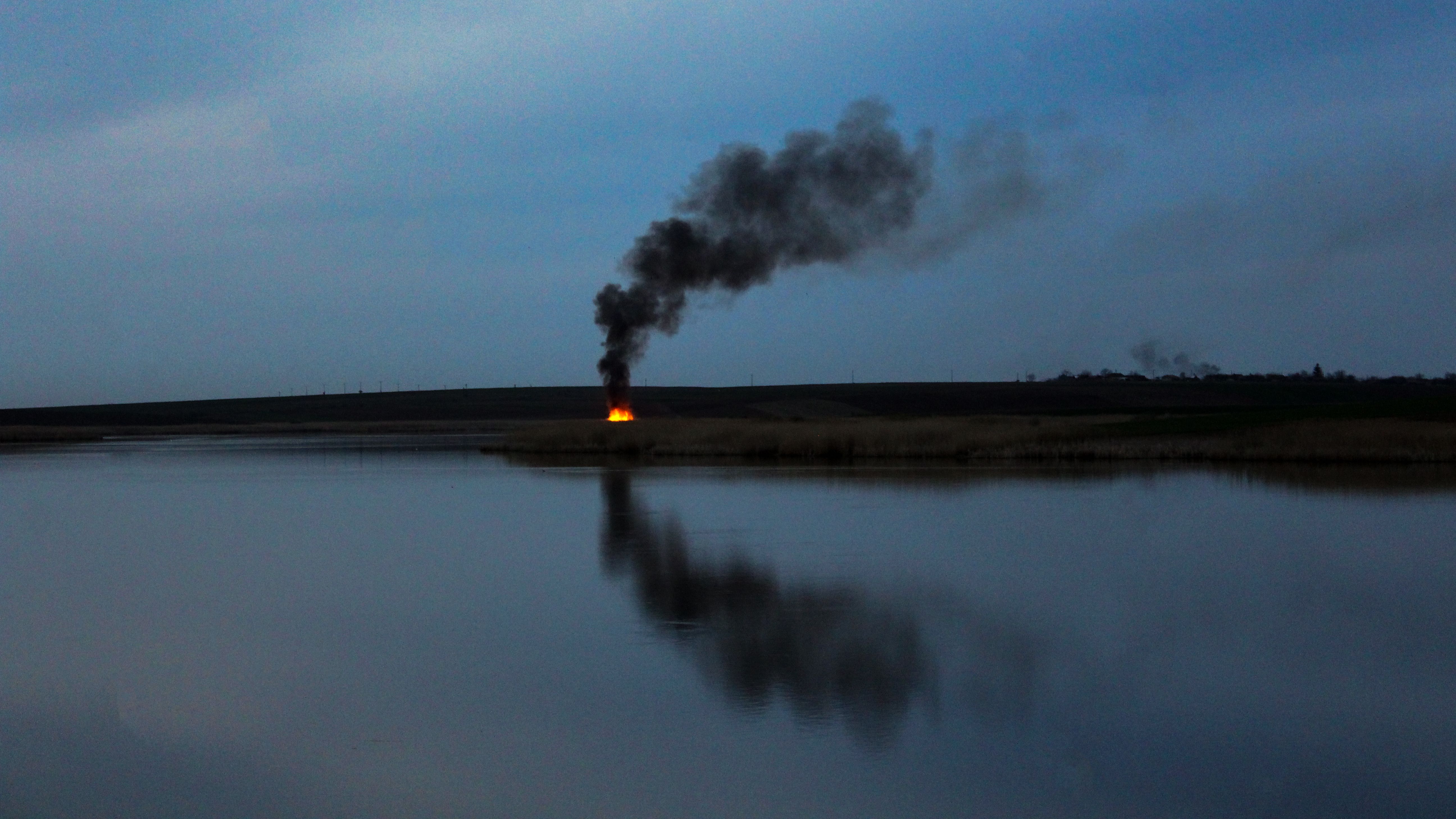 Dramatic scene of smoke rising from a fire on a lakeshore during twilight.