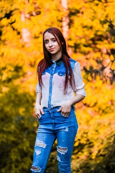 一位身着牛仔的年轻女子，自信地站在秋叶生机勃勃的景色中，散发着时尚与魅力