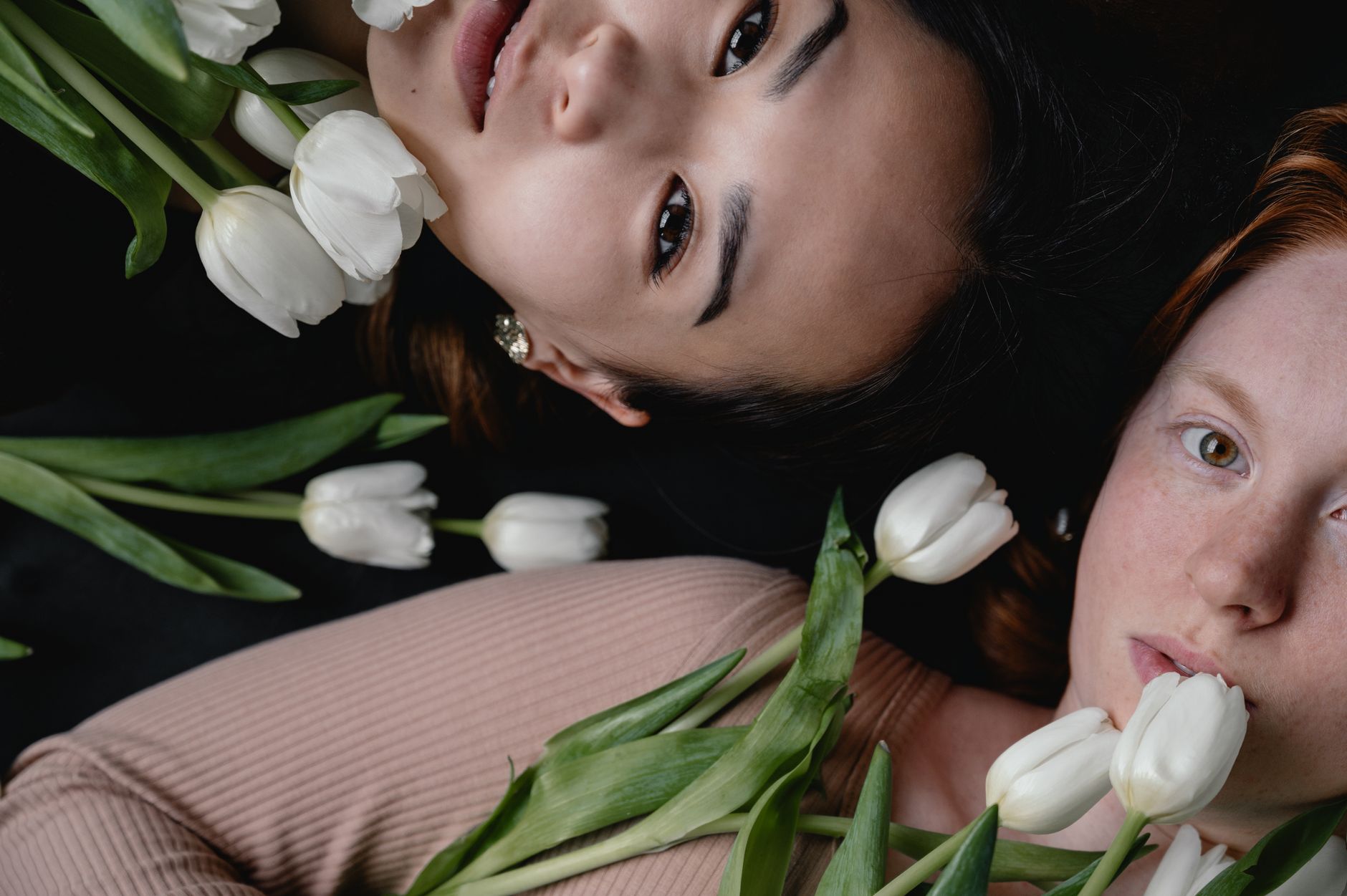 Close-up portrait of two women lying with white tulips, creating an elegant and serene composition.