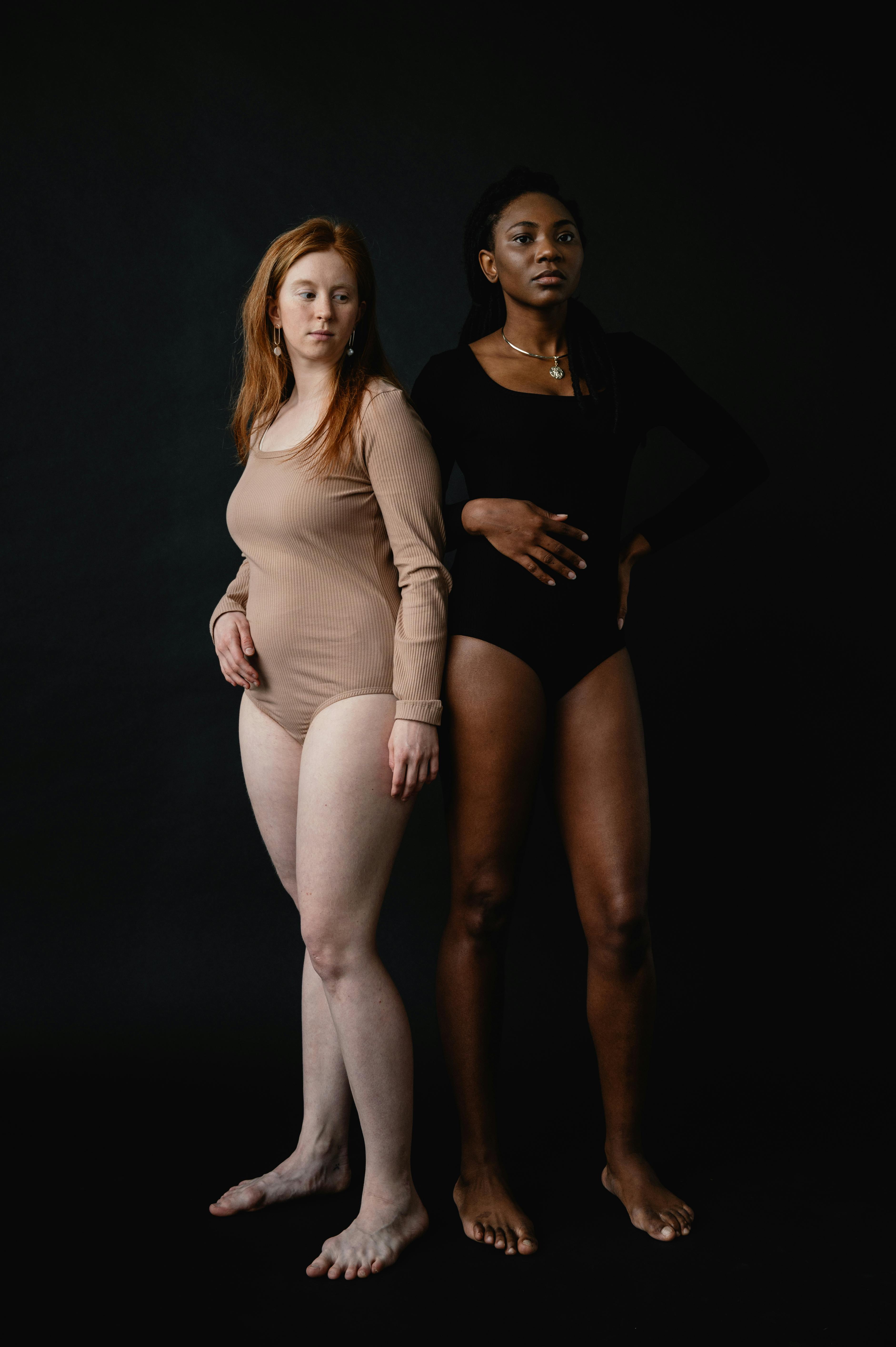 Two women in contrasting bodysuits pose confidently against a dark backdrop.