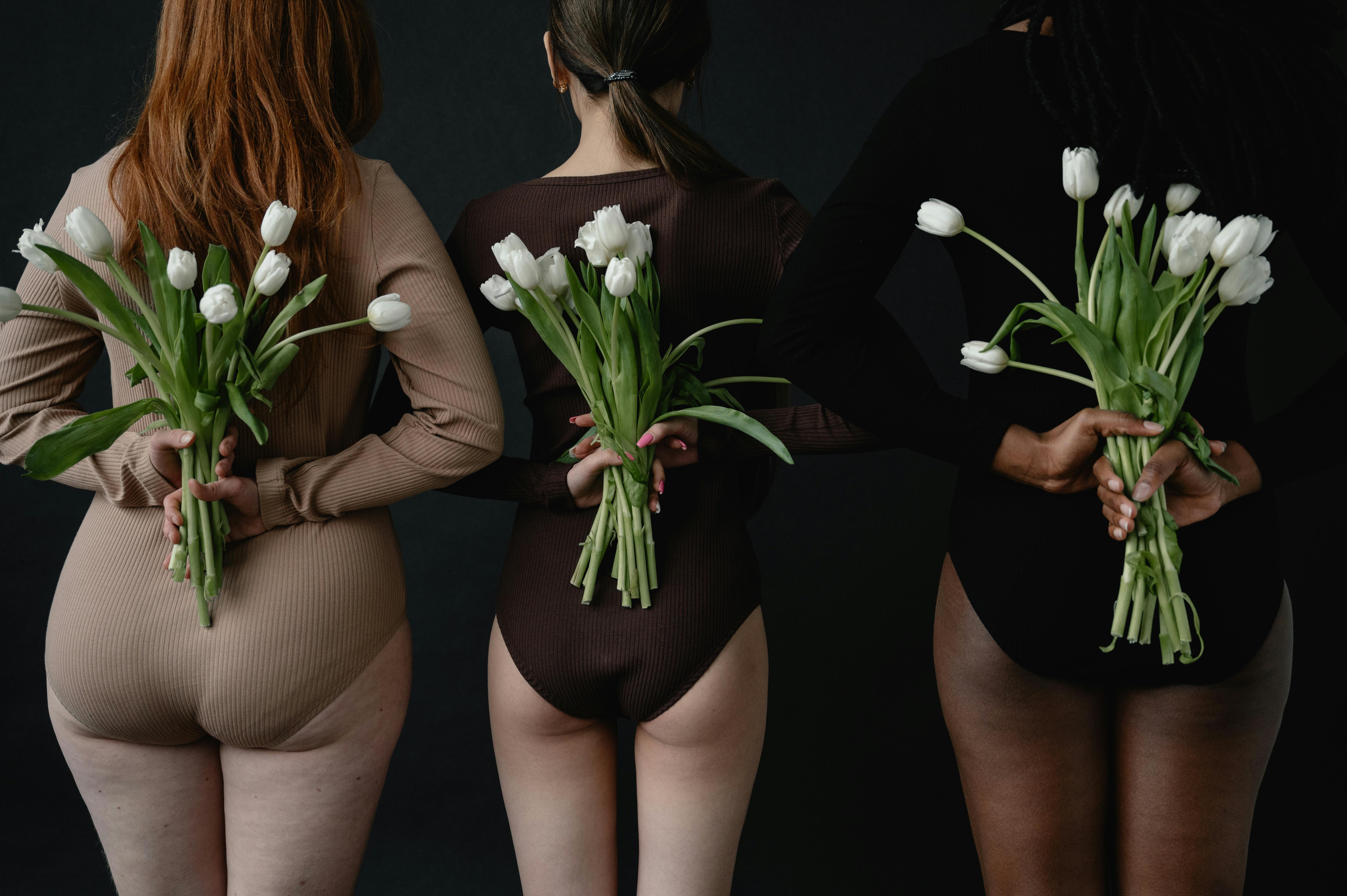 three women wearing body suits holding flowers behind their backs