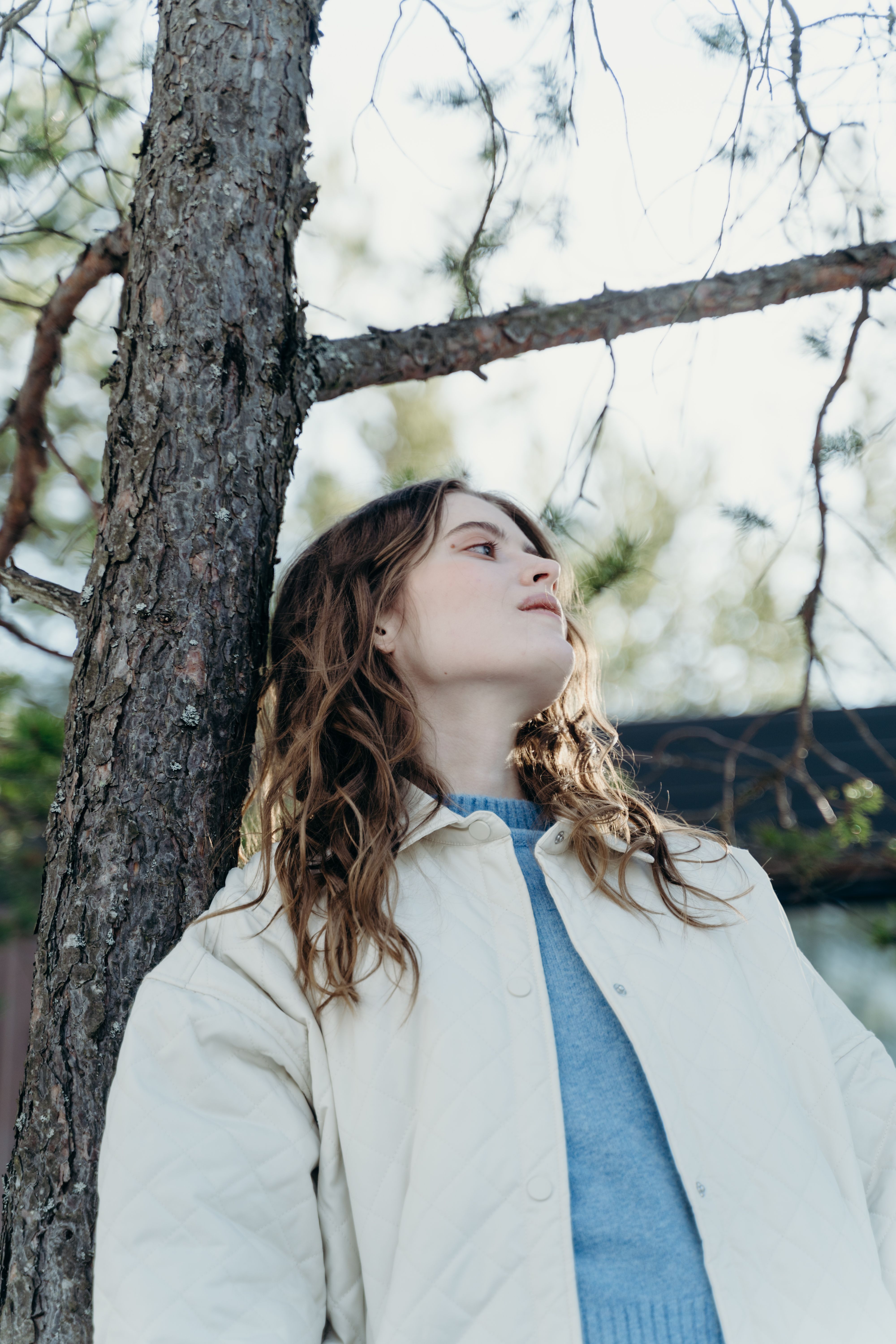 A woman in a winter jacket leaning against a tree, looking up thoughtfully.