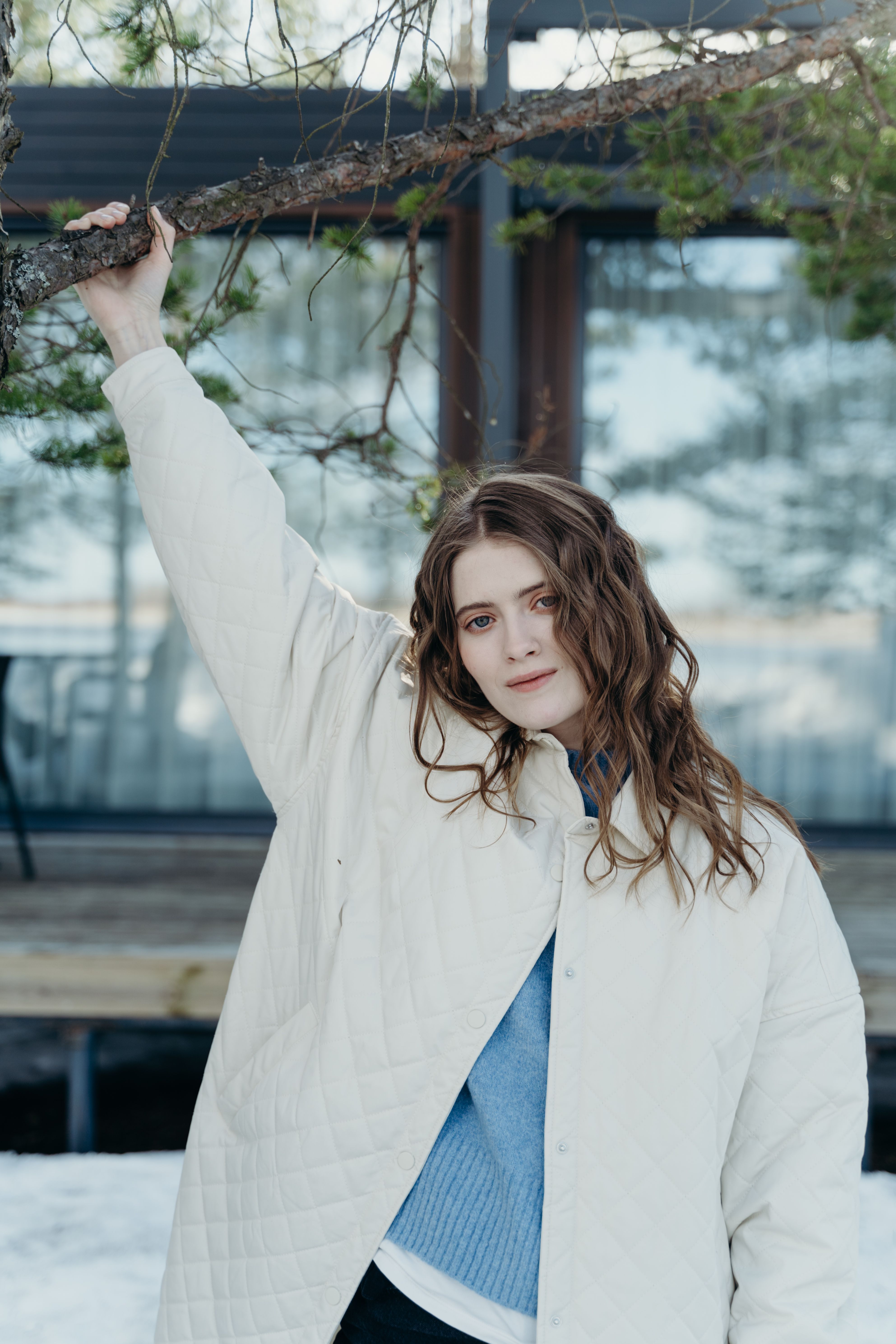 A woman in a beige winter coat stands outdoors holding a branch, exuding a calm demeanor.