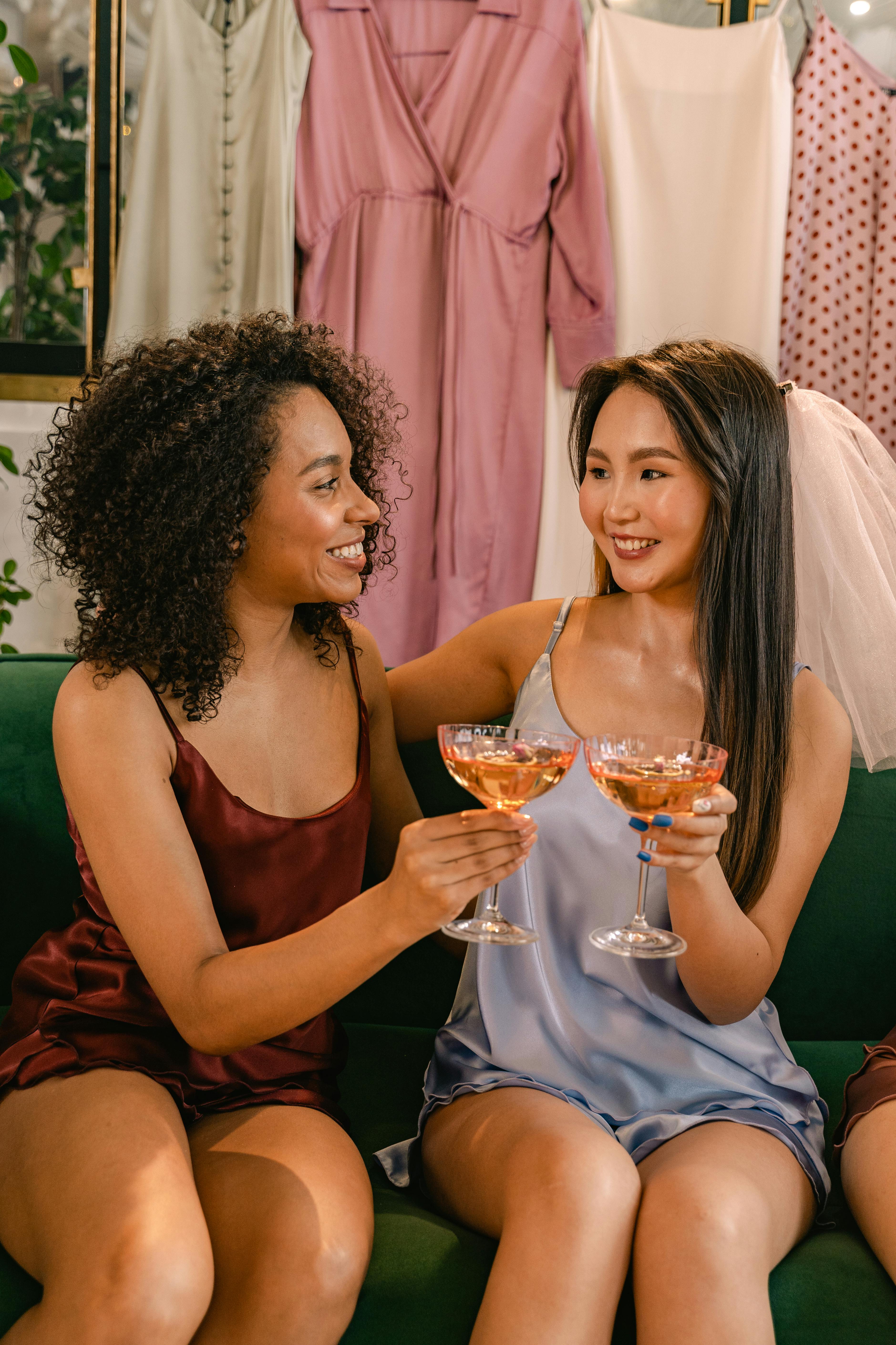 Two bridesmaids wearing satin dresses toast with cocktails, smiling and celebrating indoors.