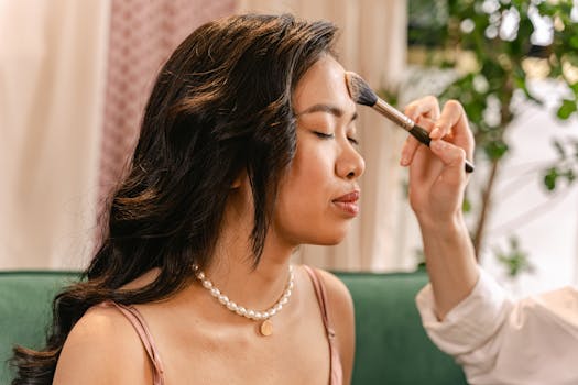Woman getting makeup applied with a brush, eyes closed, in a soft-lit indoor setting.