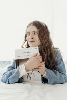 Caucasian woman in a cozy blue sweater holding a magazine on a white background indoors.