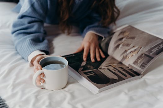 A person in a blue sweater enjoys reading a magazine in bed, sipping a hot drink.