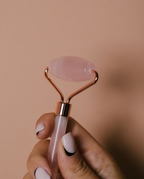 A detailed close-up of a hand holding a facial jade roller with manicured nails against a neutral background.