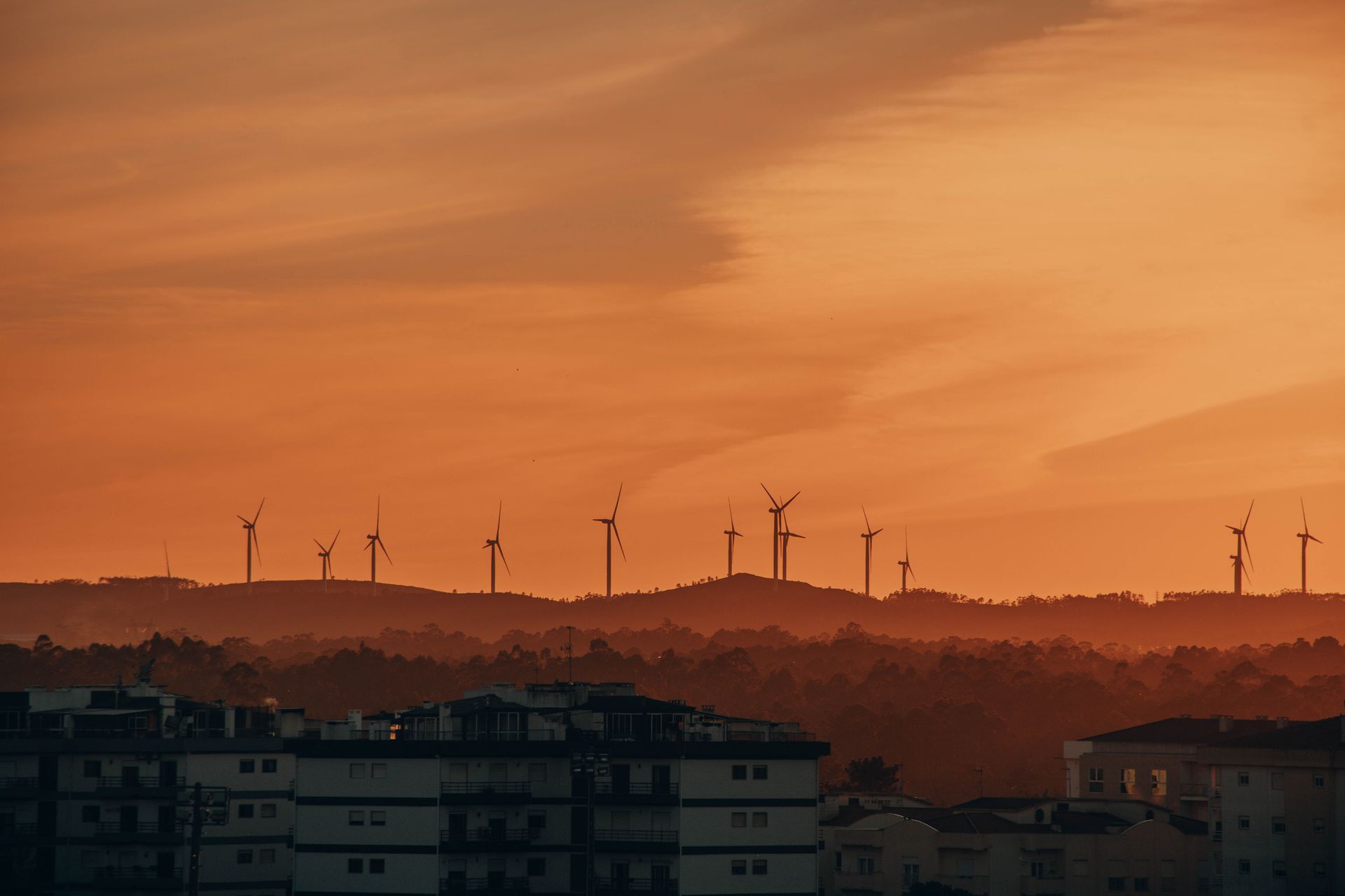 A picturesque view of wind turbines against an evening sunset sky over a silhouetted urban landscape.
