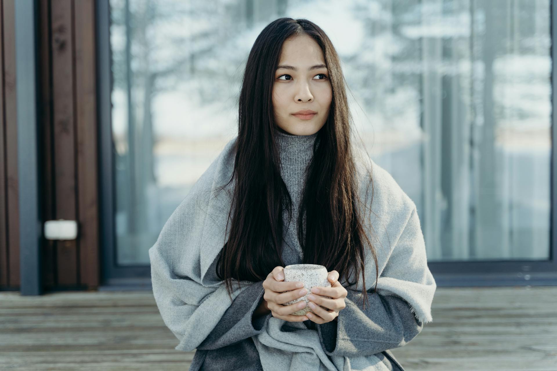 Young woman in a winter coat, holding a hot drink, sitting outside on a cold day.