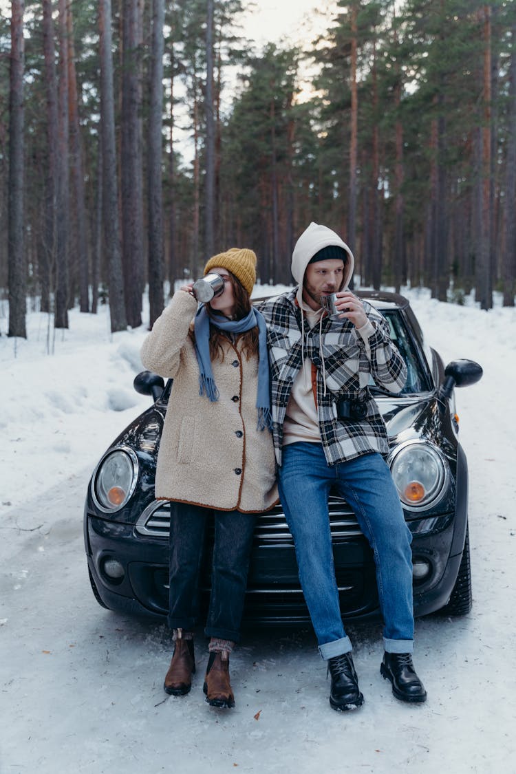 A Couple In Winter Clothes Sipping Coffee While Leaning On A Car