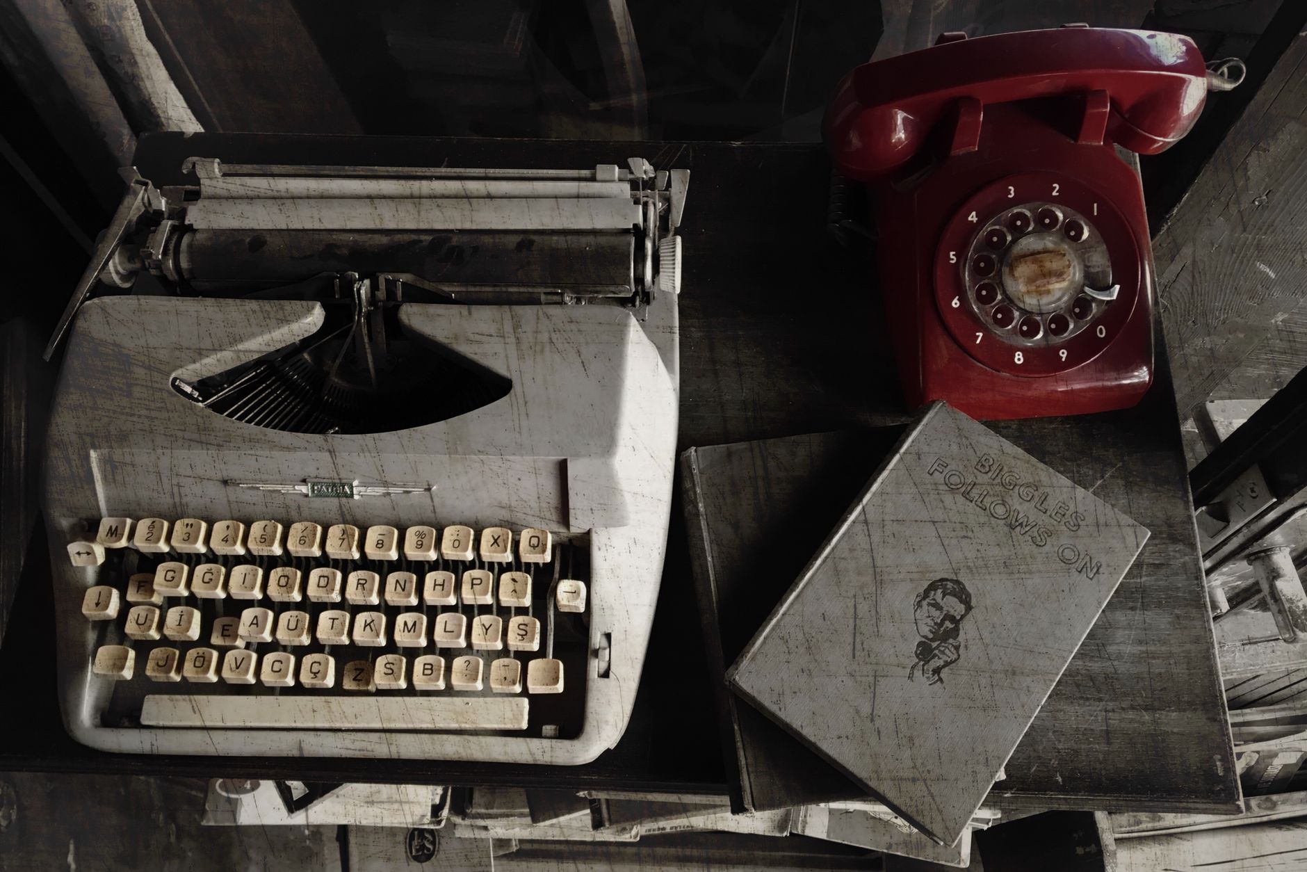 Top view of vintage typewriter, red rotary phone, and book on a wooden desk.