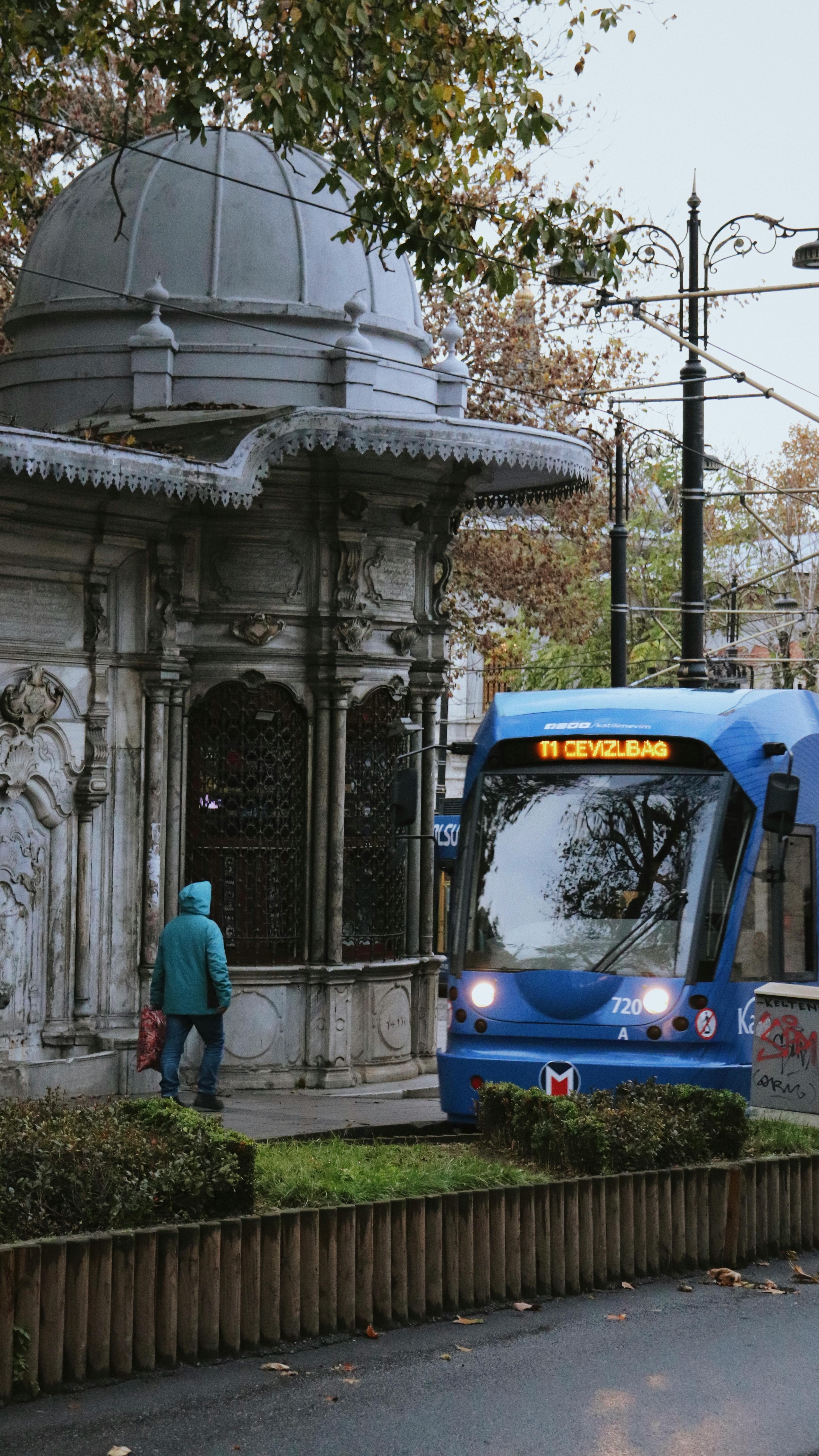 Free Tram moving past a historic stone kiosk on a rainy city day, capturing urban and transport themes. Stock Photo