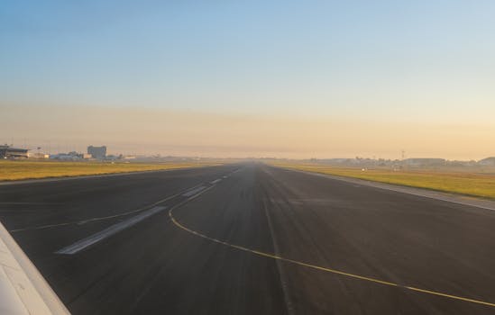 An empty airport runway at sunrise, showcasing a clear sky and distant airport buildings.