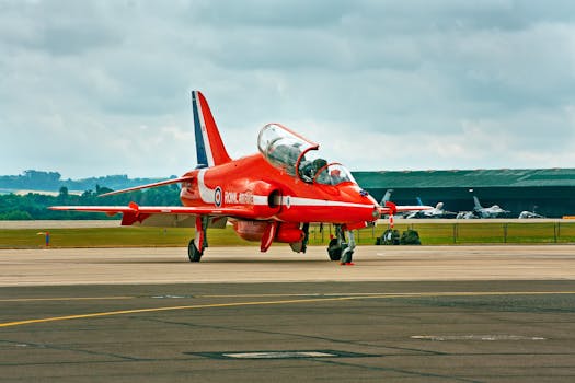 Red Arrows training jet parked on a runway in England, cloudy sky above.