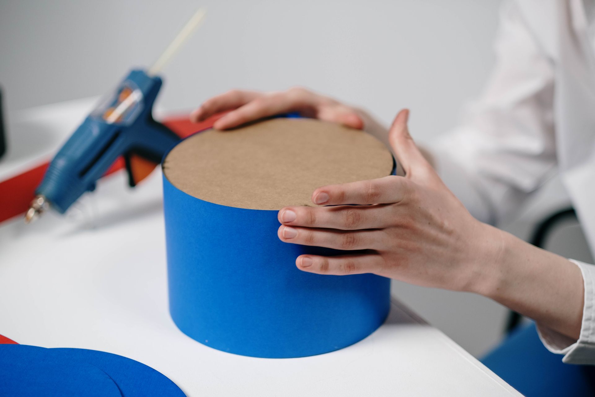 Hands crafting a blue cardboard cylinder with glue gun on a desk.