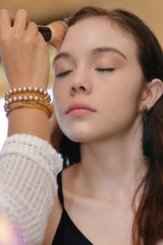 Close-up of a makeup artist applying foundation on a young woman's face indoors.