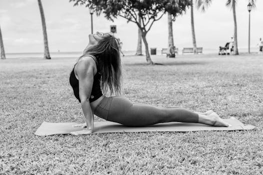 A woman in a yoga pose on a mat outdoors, expressing strength and flexibility in a grayscale image.