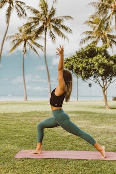 A woman performs yoga on a mat in a park, surrounded by palm trees under a clear sky.