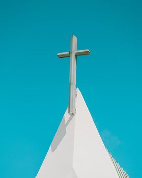 A minimalist shot of a church cross atop a triangular roof against a clear blue sky.