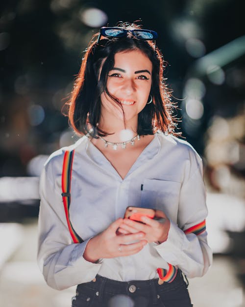 Cheerful young ethnic female with dark hair in casual clothes smiling and looking at camera while messaging on mobile phone standing on city street on sunny day