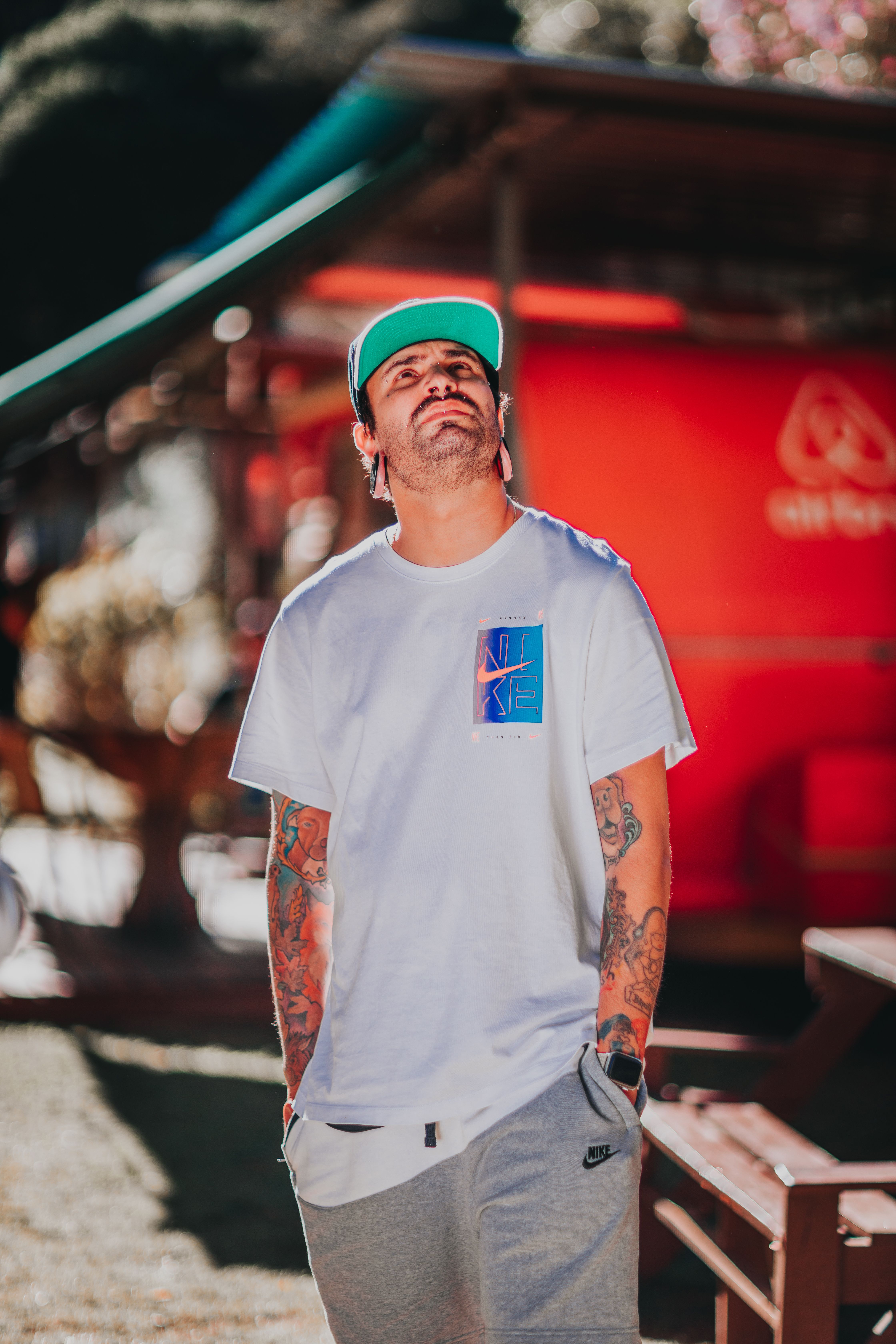Concentrated young unshaven male millennial with tattoos on arms in casual outfit and cap standing on city street with hands in pockets and looking up thoughtfully