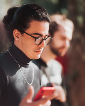 Concentrated young ethnic trendy male with long dark hair in stylish turtleneck and eyeglasses reading message on mobile phone while standing on street on sunny day