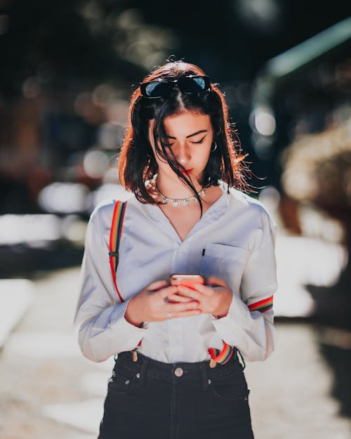 Free Trendy ethnic female teen with dark hair in casual outfit and sunglasses on head using mobile phone standing in city on sunny day Stock Photo