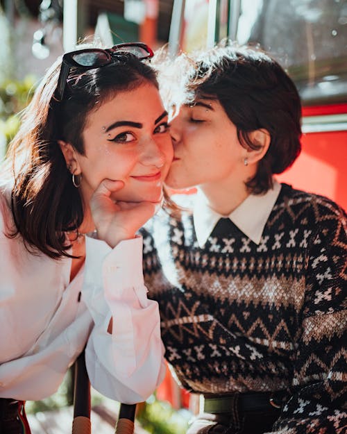 Romantic young lesbian female millennial with dark hair in casual clothes kissing cheek of beloved girlfriend with closed eyes while resting together in street cafe on sunny day