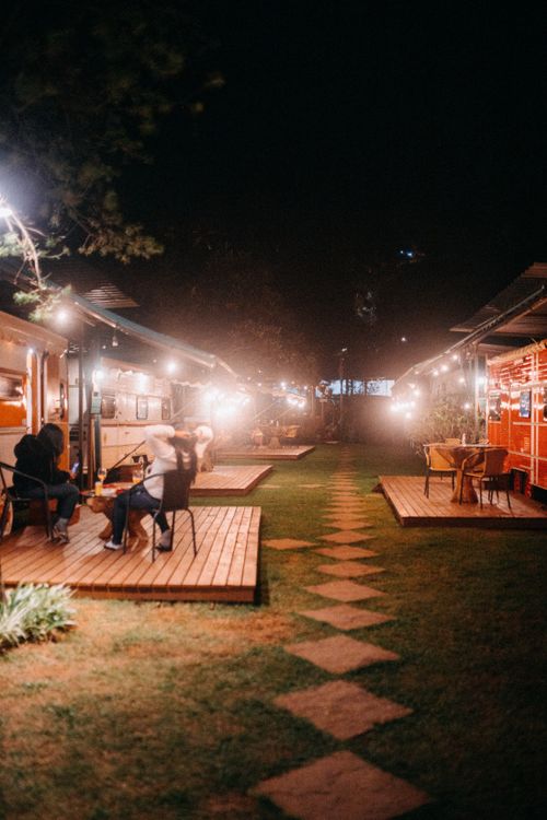 Free People chilling at table on wooden floor near white camping van with glowing streetlights spending evening in leisure Stock Photo