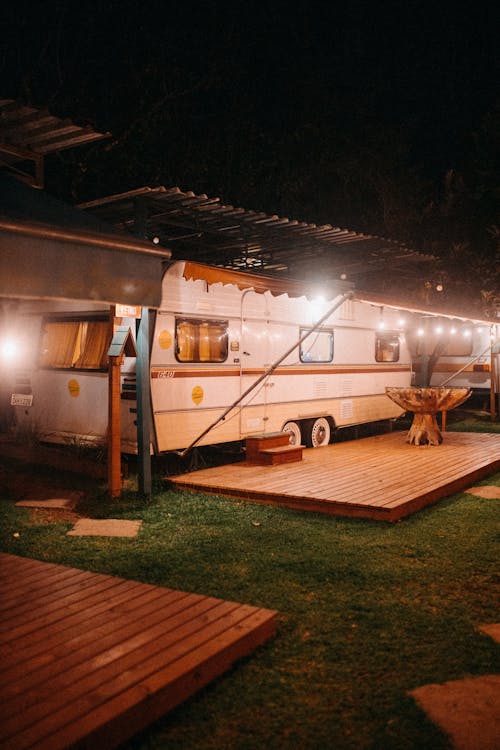 White camping trailer parked under roof with glowing garlands near wooden floor with stairs and table at night
