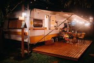 Side view of female tourist at table with alcoholic drink against camping trailer with shiny lamps at night