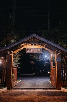 Wooden arch with gate and title on sign over asphalt roadway between trees illuminated by shiny lamps at night in campground