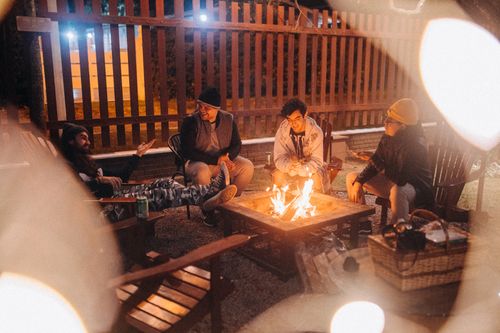 Free Male travelers speaking while sitting in armchairs against shiny flame and fence at night in campground Stock Photo