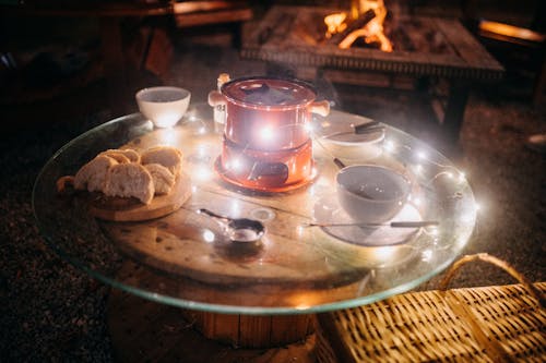 Small fondue pot and skewers with cups placed on woodn and glass table with served bread pieces for picnic in night garden