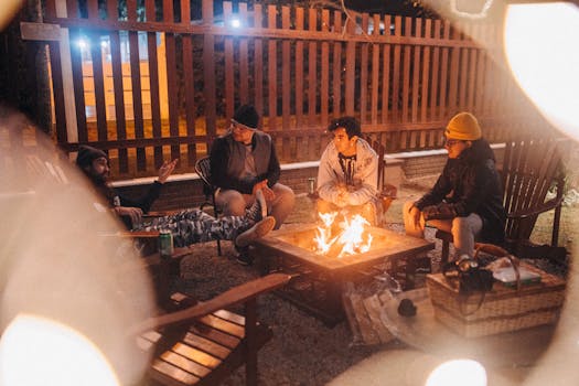 Group of friends in warm clothes talking while sitting around burning fire near fence in yard at night