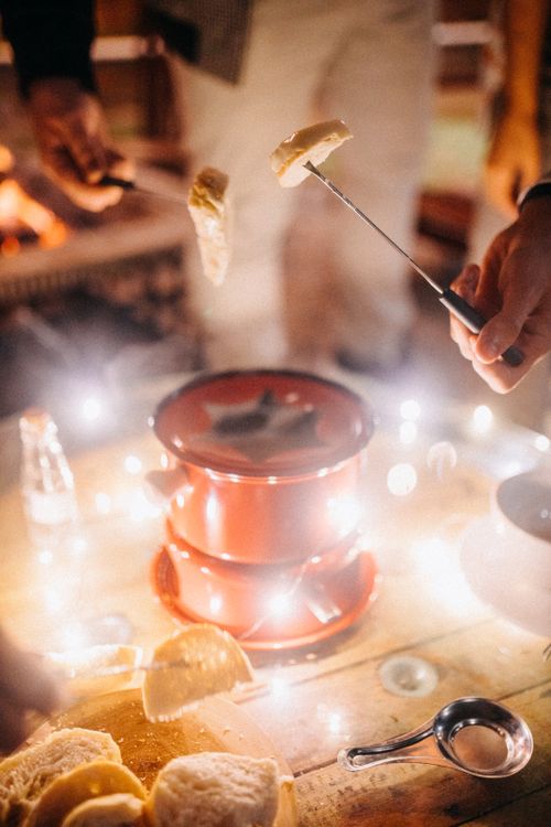 Free Crop anonymous friends melting cheese in fondue and eating bread while standing around wooden table with garland at night Stock Photo