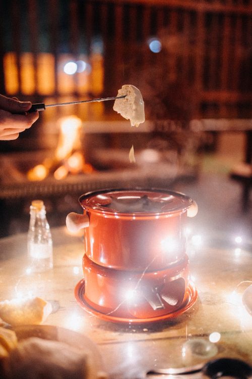 Free Crop anonymous person dipping piece of bread in melt cheese in fondue pot placed on glass table with garland in yard at night Stock Photo