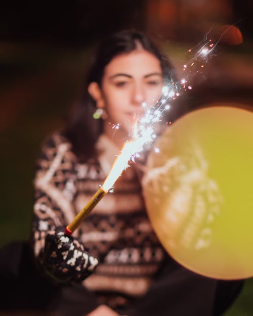 Young female in warm patterned sweater holding burning sparkler while looking at camera on street at night
