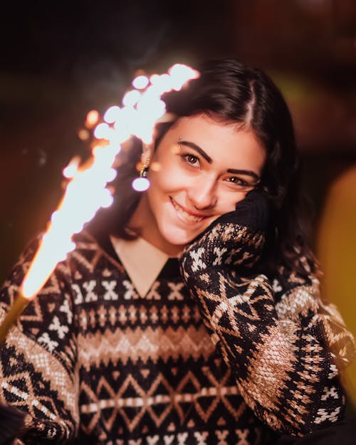 Happy young female in warm clothes smiling and holding burning Bengal light while looking at camera and standing on street at night