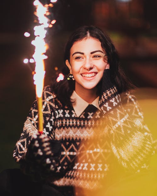 Happy young female in stylish outfit looking at burning Bengal light and smiling on blurred background of street at night