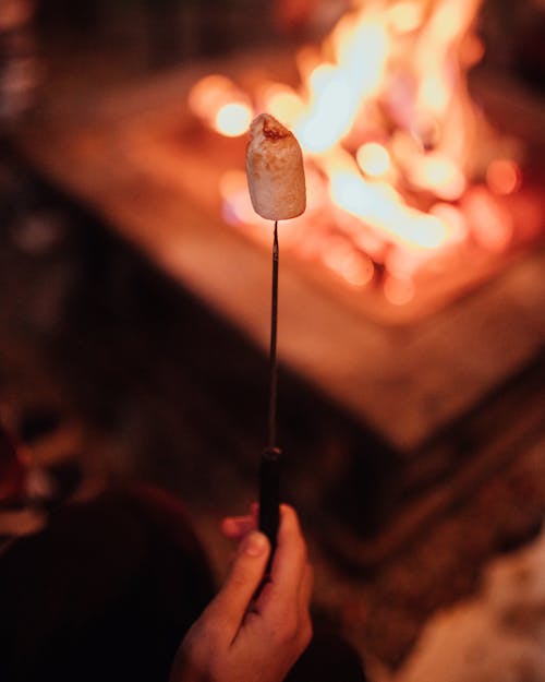 Crop anonymous person holding stick with delicious fried marshmallow near burning flame onn street in evening