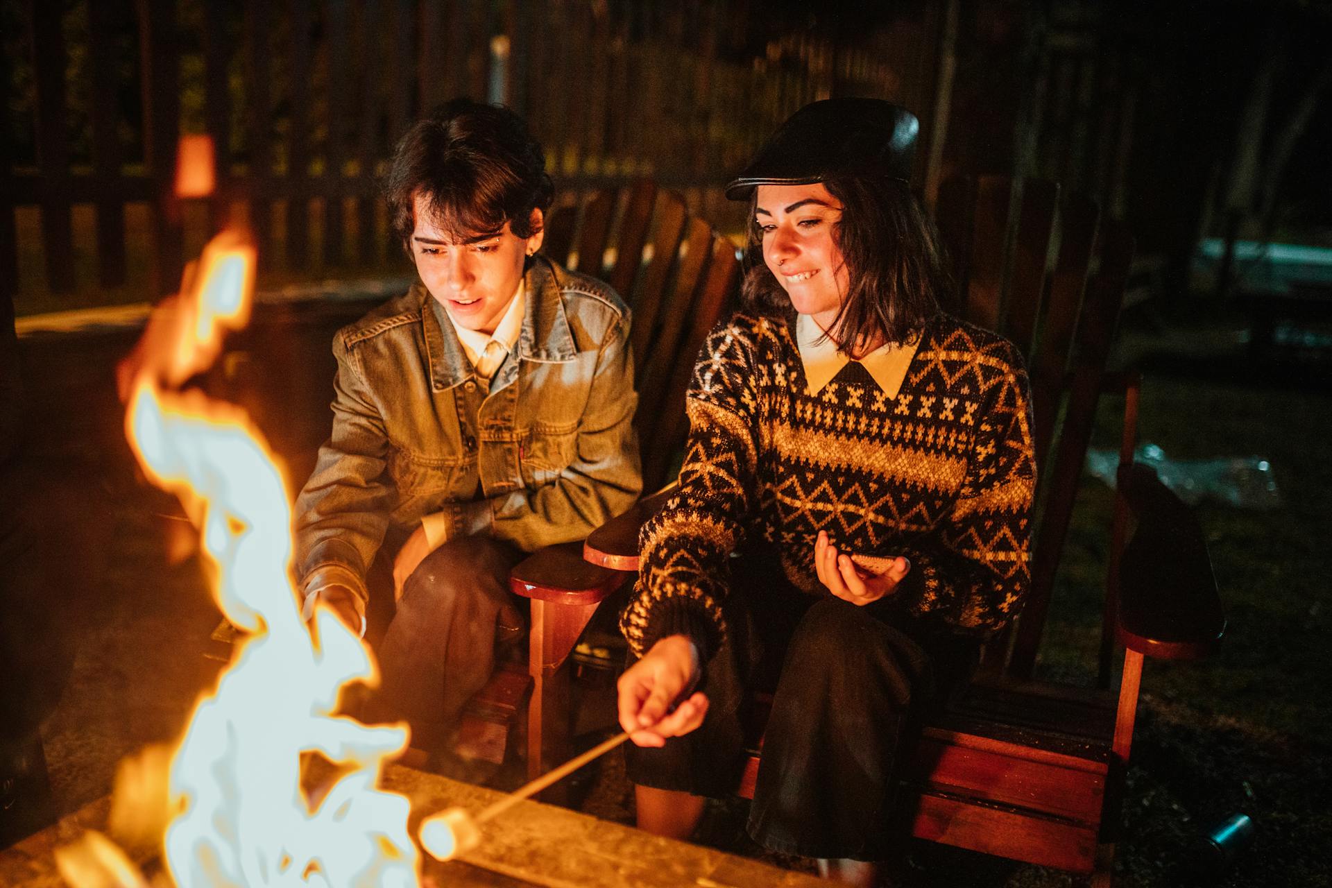 Concentrated friends sitting together in armchairs in backyard and looking at burning bonfire while roasting marshmallow at night