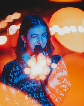 Smiling young woman in patterned sweater holding burning sparkler and looking at camera while standing on city street at night
