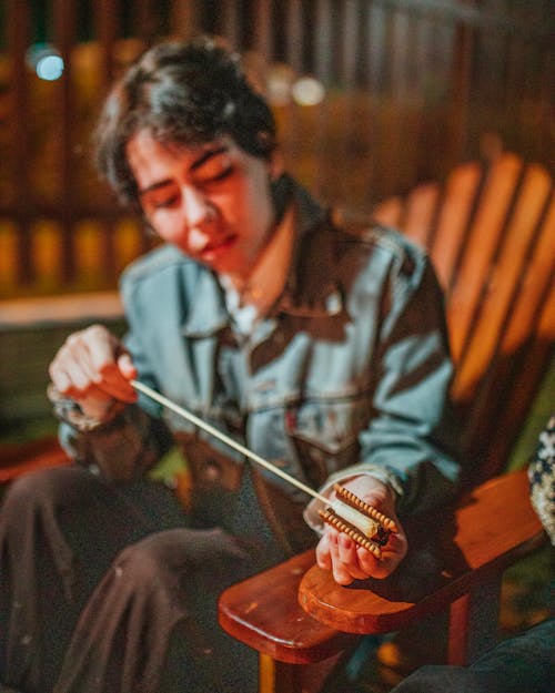 Young female putting stick with marshmallow between graham crackers while preparing campfire treat at dusk