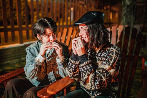 Smiling female friends looking at each other while eating tasty campfire treats in armchairs against fence in evening